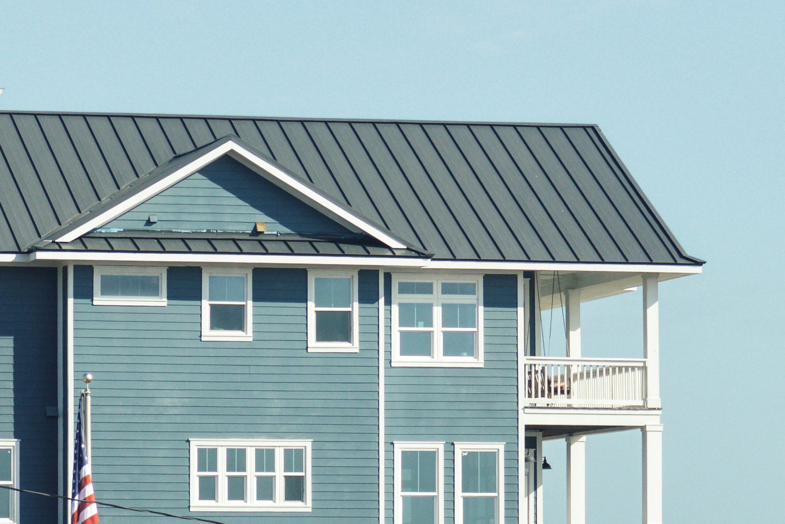 Blue two-story house with a grey metal roof and american flag displayed on a clear day.