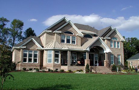 Two-story brick house with large windows, a pointed roof, a front porch, and surrounding greenery under a blue sky.