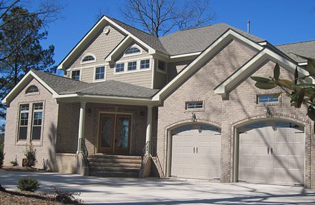 A two-story suburban home with a beige brick exterior, twin garage doors, and a large front entrance under a clear blue sky.