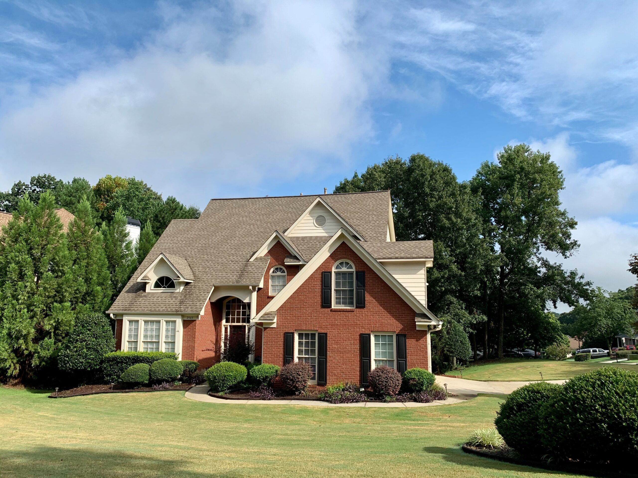 A red brick two-story house with a brown shingled roof, surrounded by a well-manicured lawn and trees under a clear blue sky.
