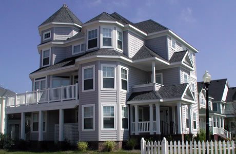 Large gray three-story house with white trim, balconies, and a fence under blue sky.